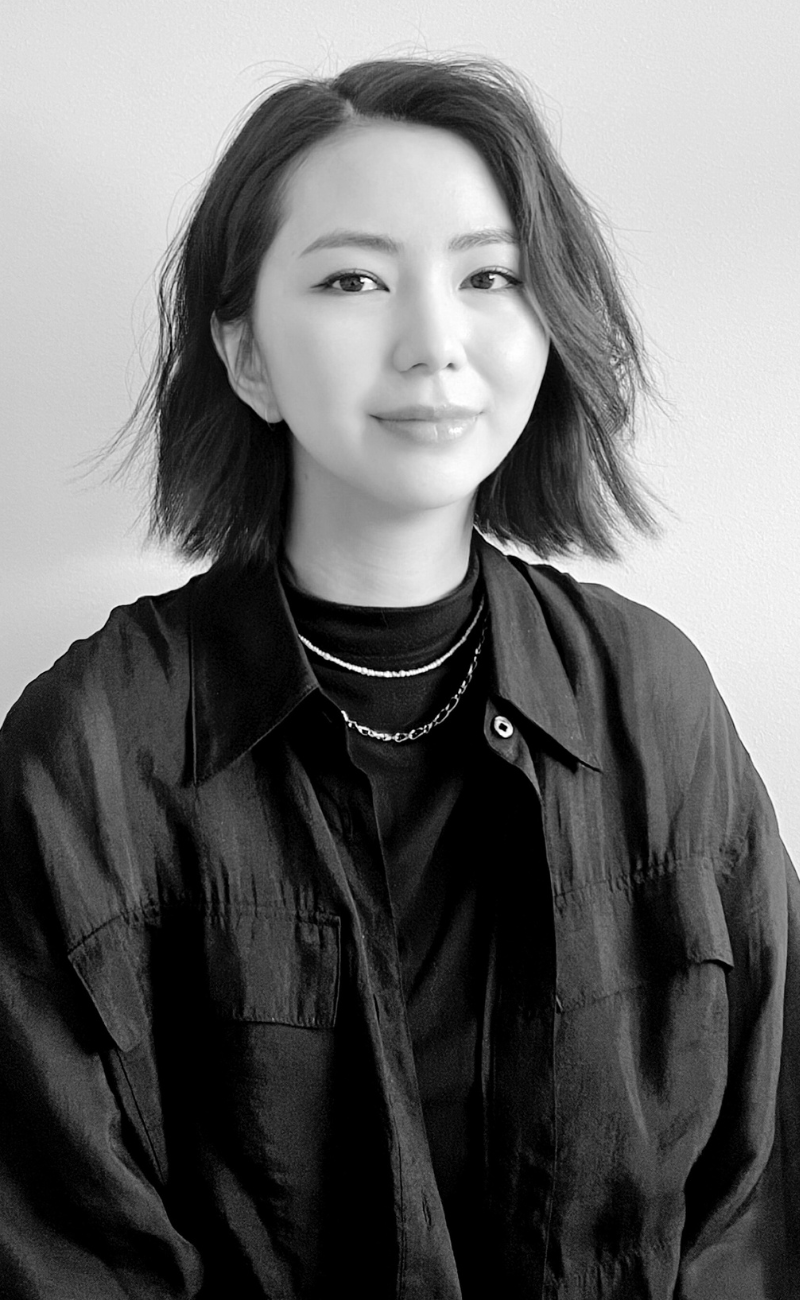 Black and white portrait of a woman with shoulder-length dark hair, wearing a white top. She has a calm and confident expression, looking directly at the camera. The background features a textured brick wall.