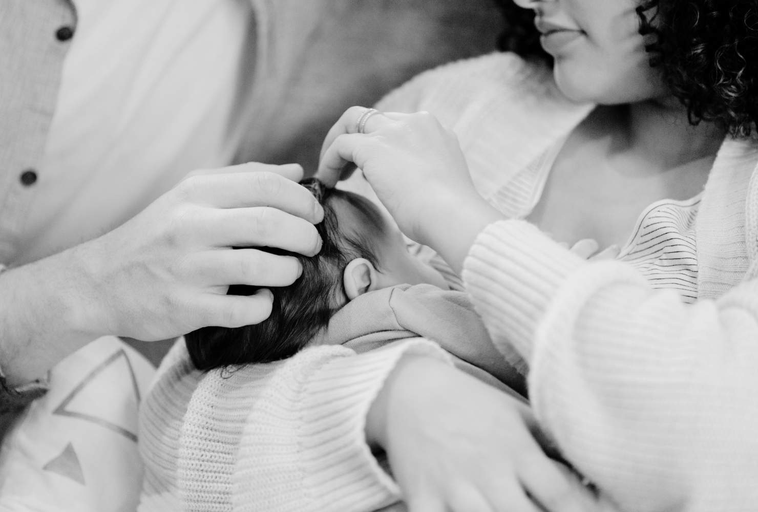 Black and white photo of a man with short curly hair and a beard, reclining on a modern couch while working on his laptop. He is wearing a relaxed white button-up shirt and dark pants. A patterned pillow rests behind him, and the setting has a minimalist, contemporary feel.