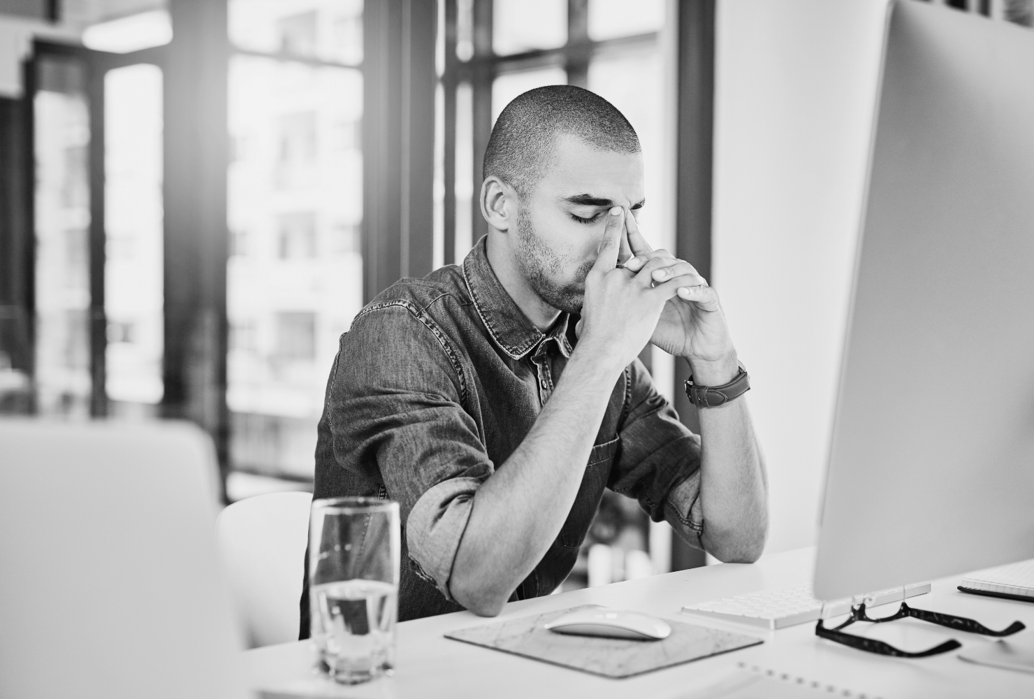 Black and white photo of a man with short curly hair and a beard, reclining on a modern couch while working on his laptop. He is wearing a relaxed white button-up shirt and dark pants. A patterned pillow rests behind him, and the setting has a minimalist, contemporary feel.
