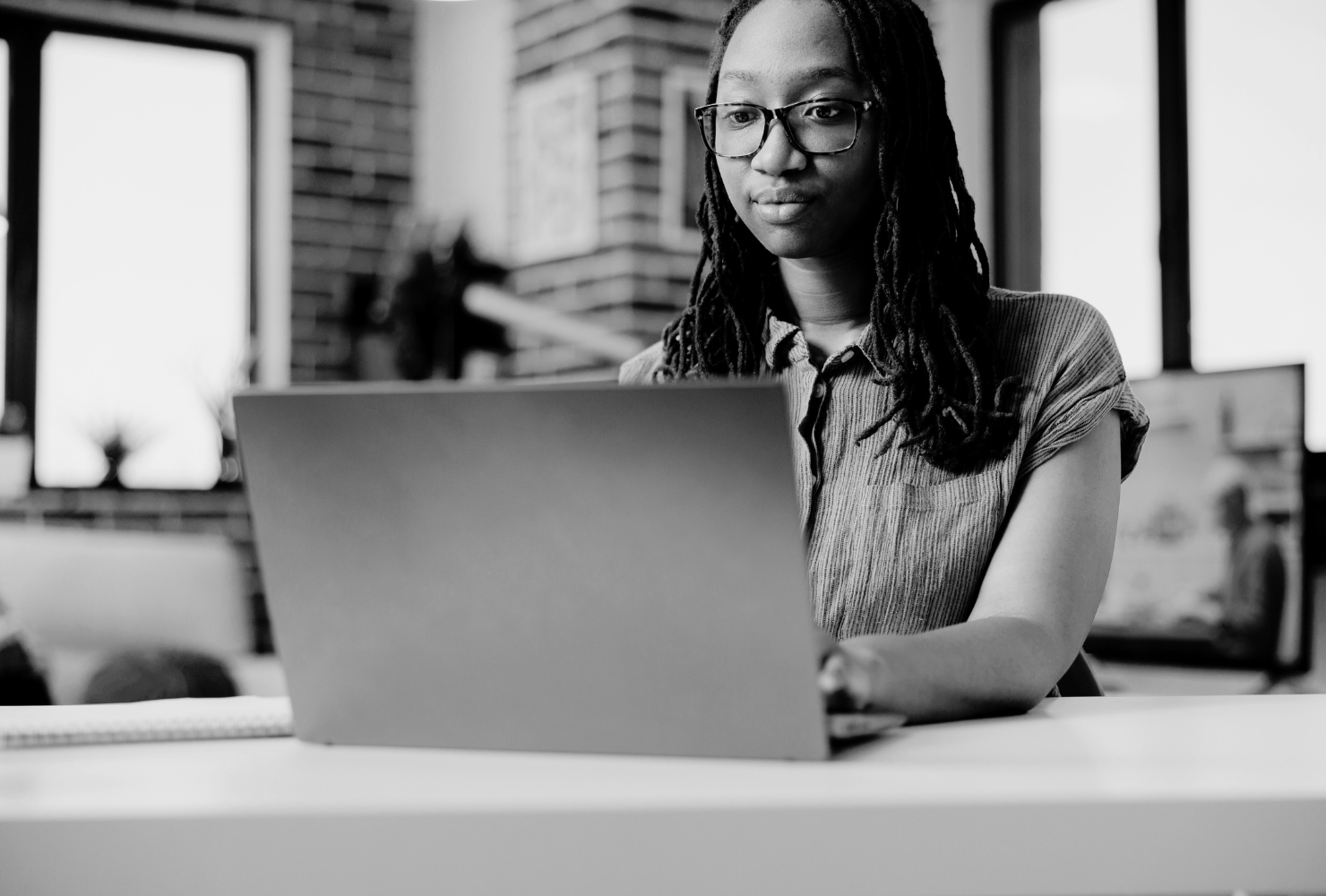 A black and white image of a couple sitting closely together on a gray couch focused on a laptop screen. The woman, with long dark hair, is wearing a cozy green cardigan over a beige top and is pointing at the screen. The man, with a beard and his hair tied in a bun, is wearing a dark green cable-knit sweater and bracelets on his wrist. The room is softly lit, with a floor lamp and decorative pillows adding warmth to the space.