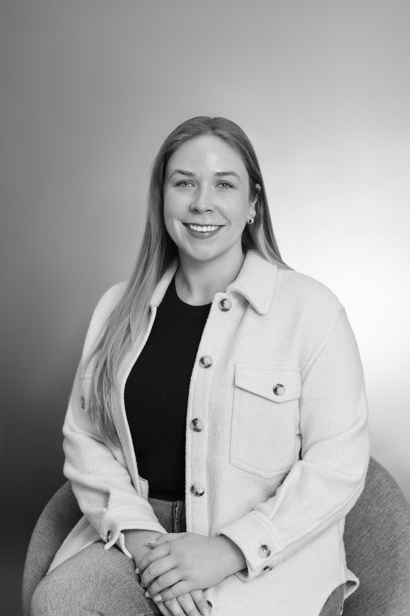 Black and white portrait of a woman with shoulder-length dark hair, wearing a white top. She has a calm and confident expression, looking directly at the camera. The background features a textured brick wall.