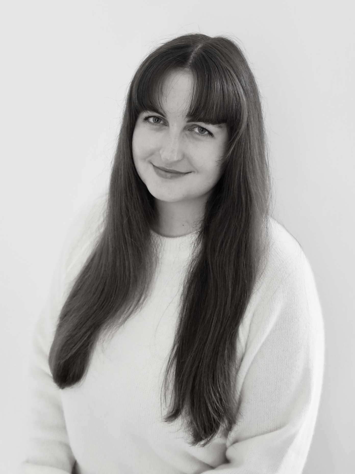 Black and white portrait of a smiling woman with long, wavy blonde hair, wearing a black top and a delicate necklace. She is looking directly at the camera with a warm and approachable expression. The background is minimal and neutral.