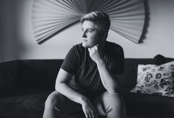 A black and white image of a couple sitting closely together on a gray couch focused on a laptop screen. The woman, with long dark hair, is wearing a cozy green cardigan over a beige top and is pointing at the screen. The man, with a beard and his hair tied in a bun, is wearing a dark green cable-knit sweater and bracelets on his wrist. The room is softly lit, with a floor lamp and decorative pillows adding warmth to the space.